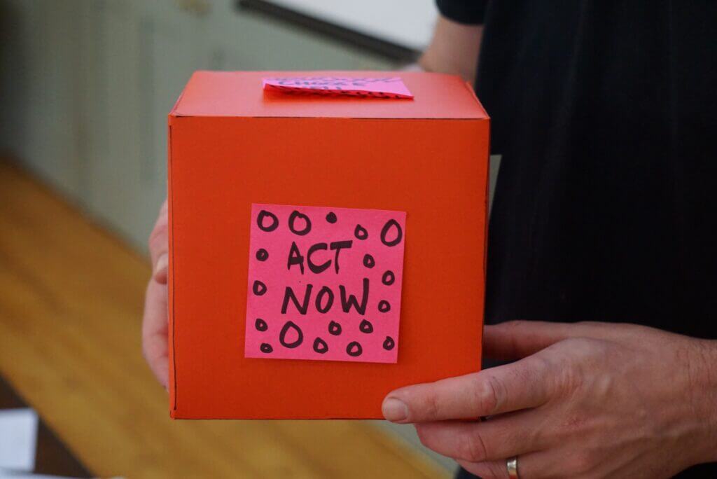 A photo of a person holding a red box with a pink sticky note reading "Act Now."