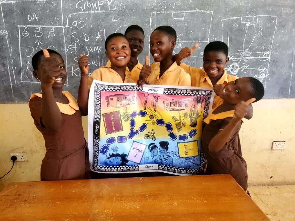 A group of smiling children standing in front of a chalkboard. The children are wearing yellow school uniforms and appear to be enthusiastic and engaged. The bright, vibrant expressions on the children's faces suggest a positive, collaborative learning environment.