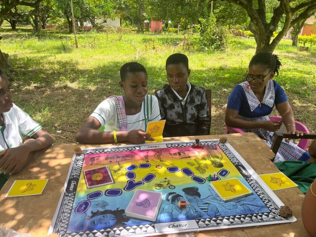 A group of children sitting around a table outdoors in a lush, green environment. The children have focused expressions as they interact with the game. The natural setting, with trees and greenery in the background, creates a pleasant, peaceful atmosphere for their activity.